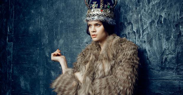 woman wearing a fur coat and a royal crown leaning against a grey slate wall